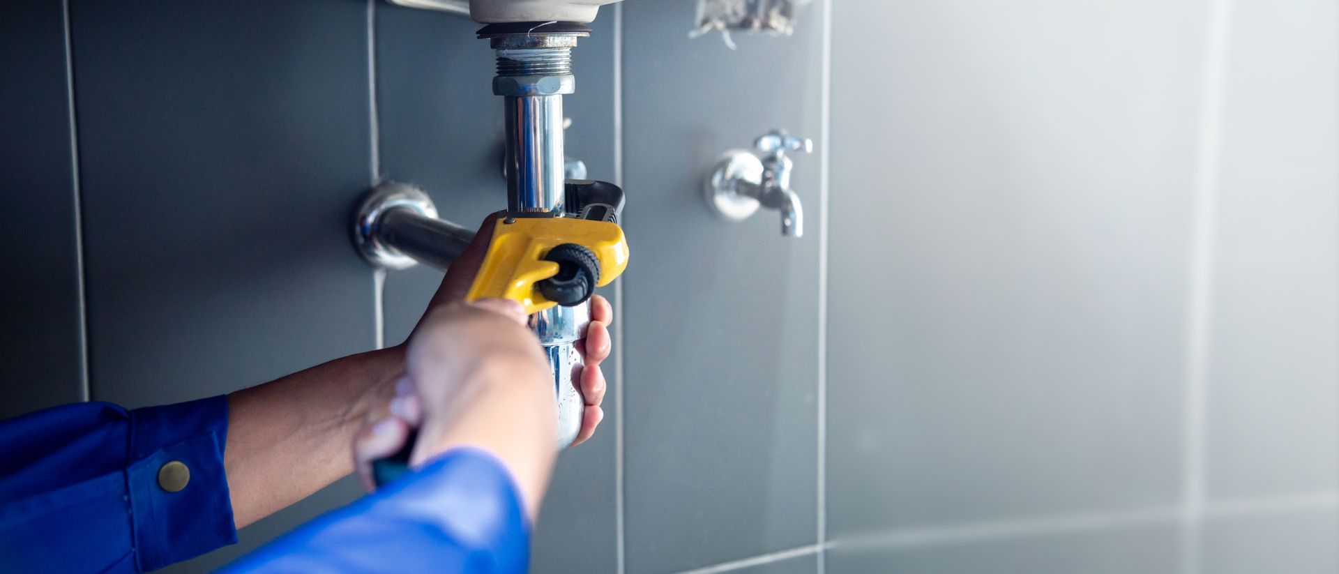 Plumber tightening a sink drain pipe connection with a pipe wrench beneath a bathroom sink. Plumber tightening a sink drain pipe connection with a pipe wrench beneath a bathroom sink.