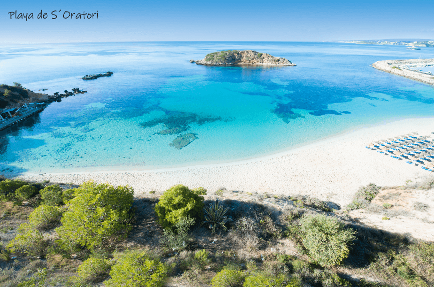 Vista aérea de una bahía azul brillante con una playa de arena, una pequeña isla y agua cristalina.