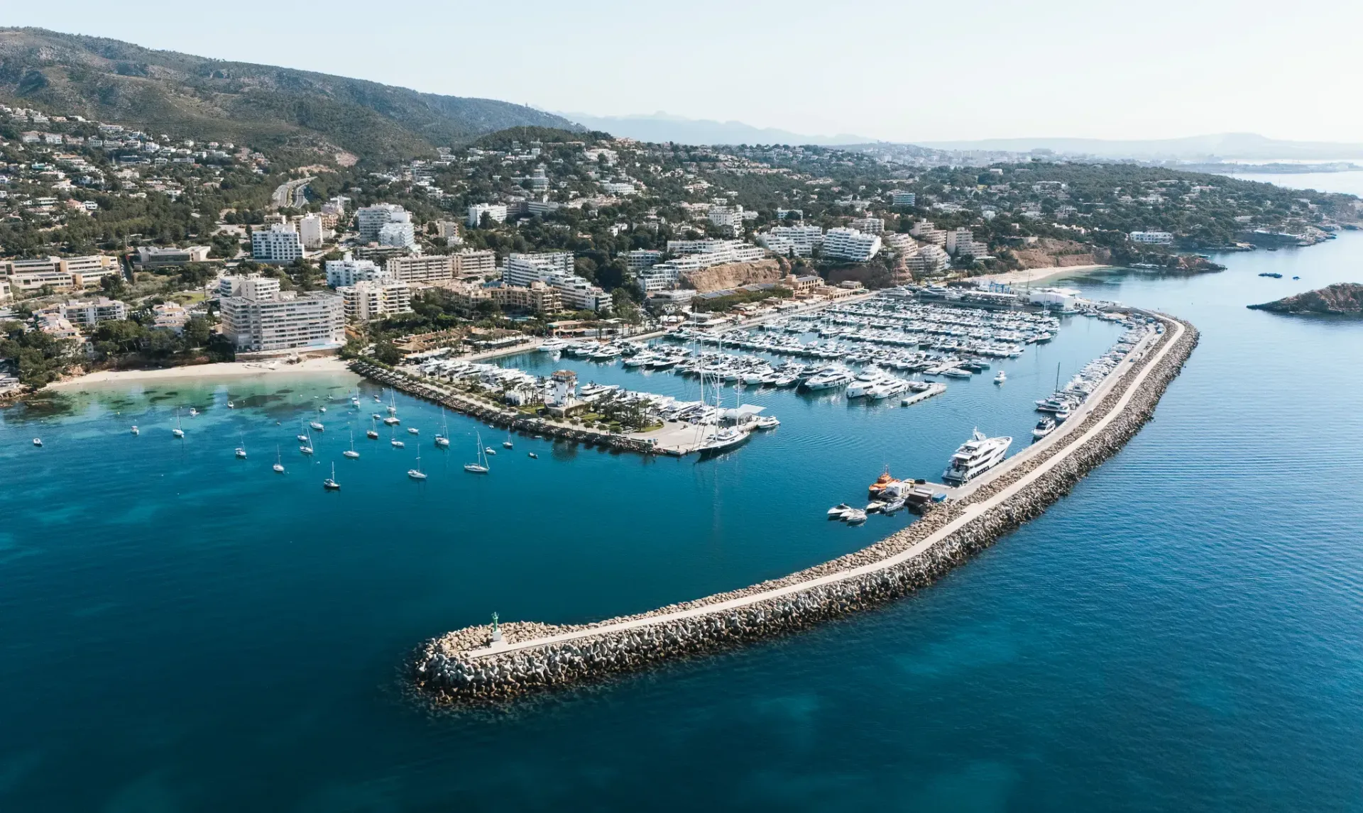 Vista aérea de un puerto deportivo con muchos barcos atracados, un largo rompeolas y una ciudad costera respaldada por una ladera cubierta de árboles.