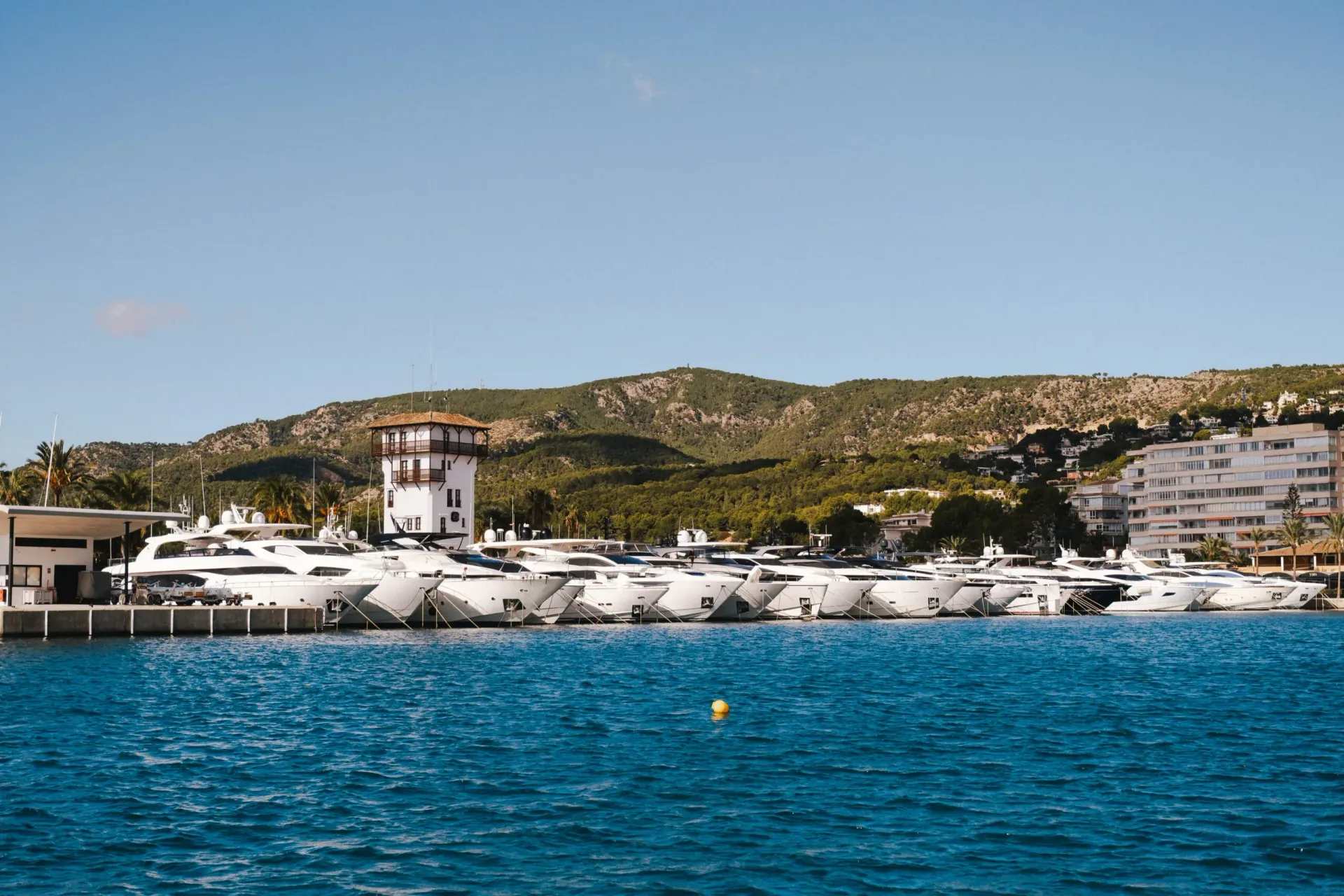 Barcos atracados en un Puerto Portals con una torre y edificios sobre un fondo de colinas verdes y un cielo azul.