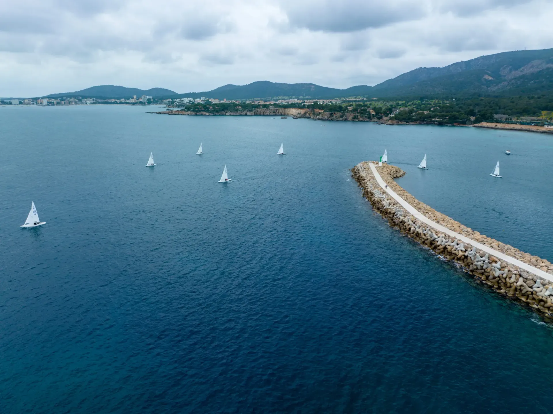 Veleros en aguas azules, un muelle rocoso y montañas verdes bajo un cielo nublado.