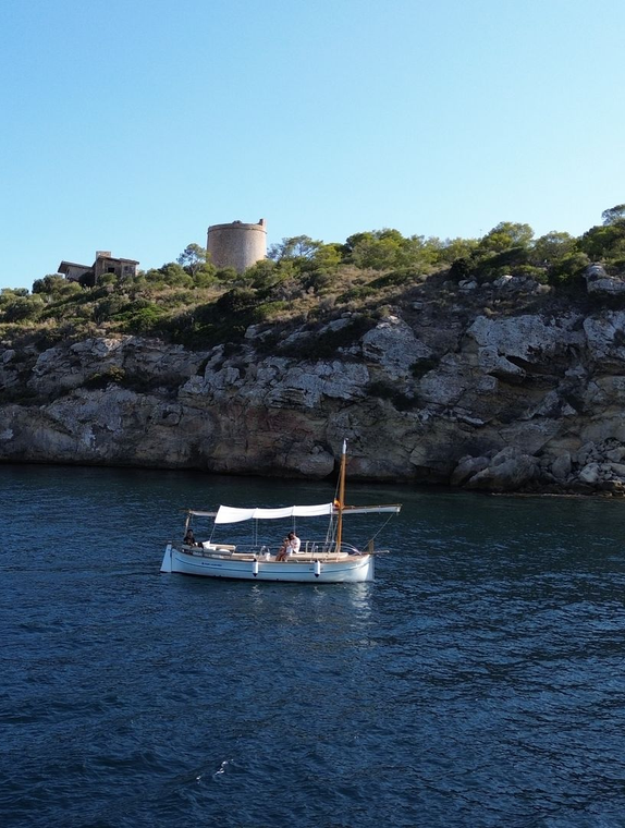 Velero en aguas azules cerca de un acantilado rocoso con una torre de piedra redonda, bajo un cielo azul claro.