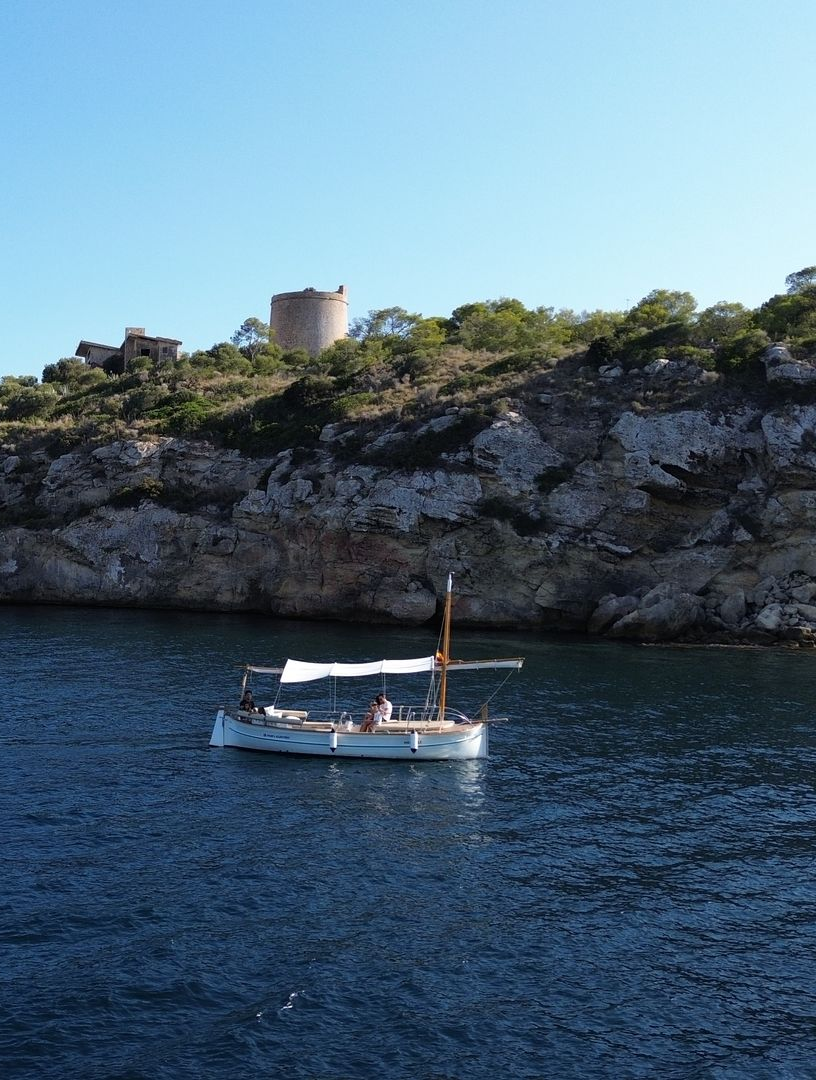 Velero en aguas azules, cerca de una costa rocosa con una torre de piedra redonda bajo un cielo despejado.