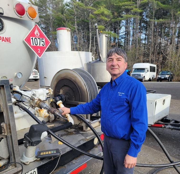 A person in a blue button-down shirt stands next to a propane truck, holding the fueling hose.
