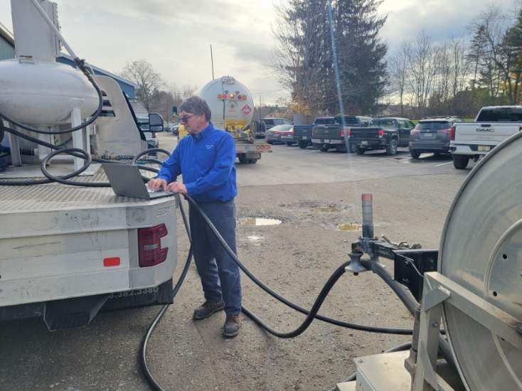 A technician in a blue shirt works on a laptop while standing next to a large utility truck at an outdoor parking lot.