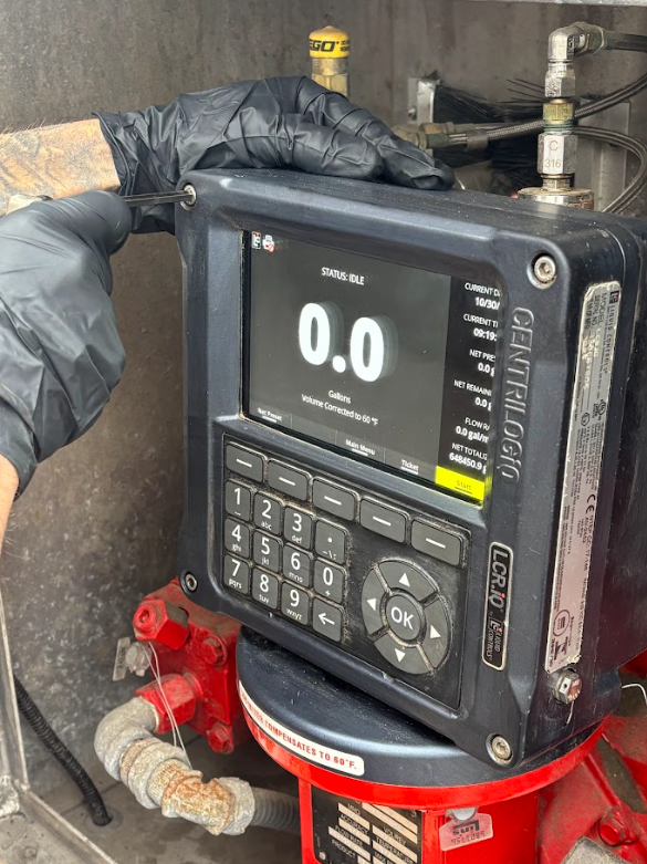 Gloved hands operate a black digital flow meter controller with a screen displaying 