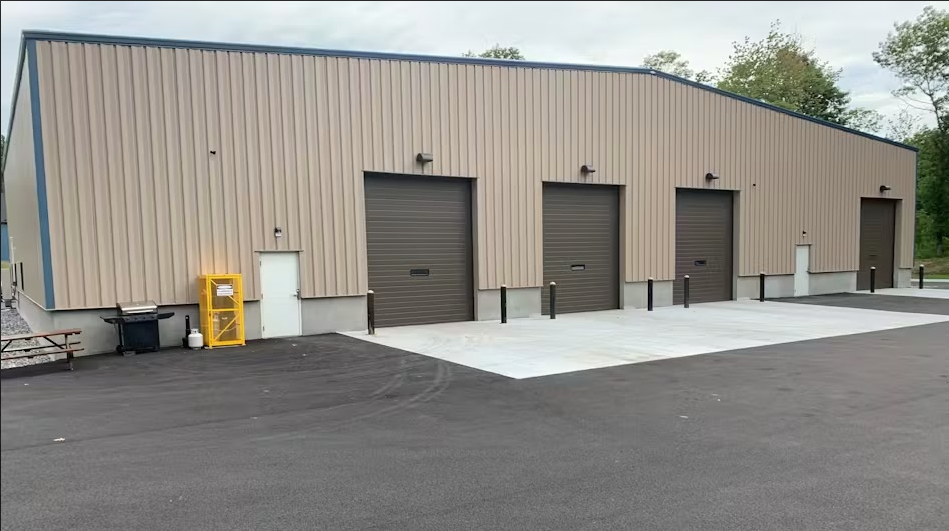 A single-story metal warehouse building with four dark-toned roll-up doors, a white entry door, and a paved lot.