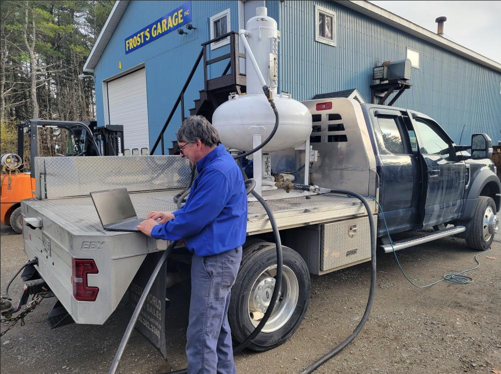 A person in a blue shirt uses a laptop on the back of a utility truck parked outside a garage.