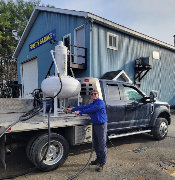 A person in a blue shirt stands next to a truck with a large, white tank, parked in front of a blue building.