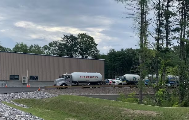A line of parked tanker trucks next to a large industrial building on a grassy lot with trees under a cloudy sky.