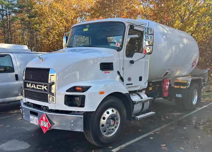 A white Mack tanker truck parked in an outdoor lot with autumn trees in the background.
