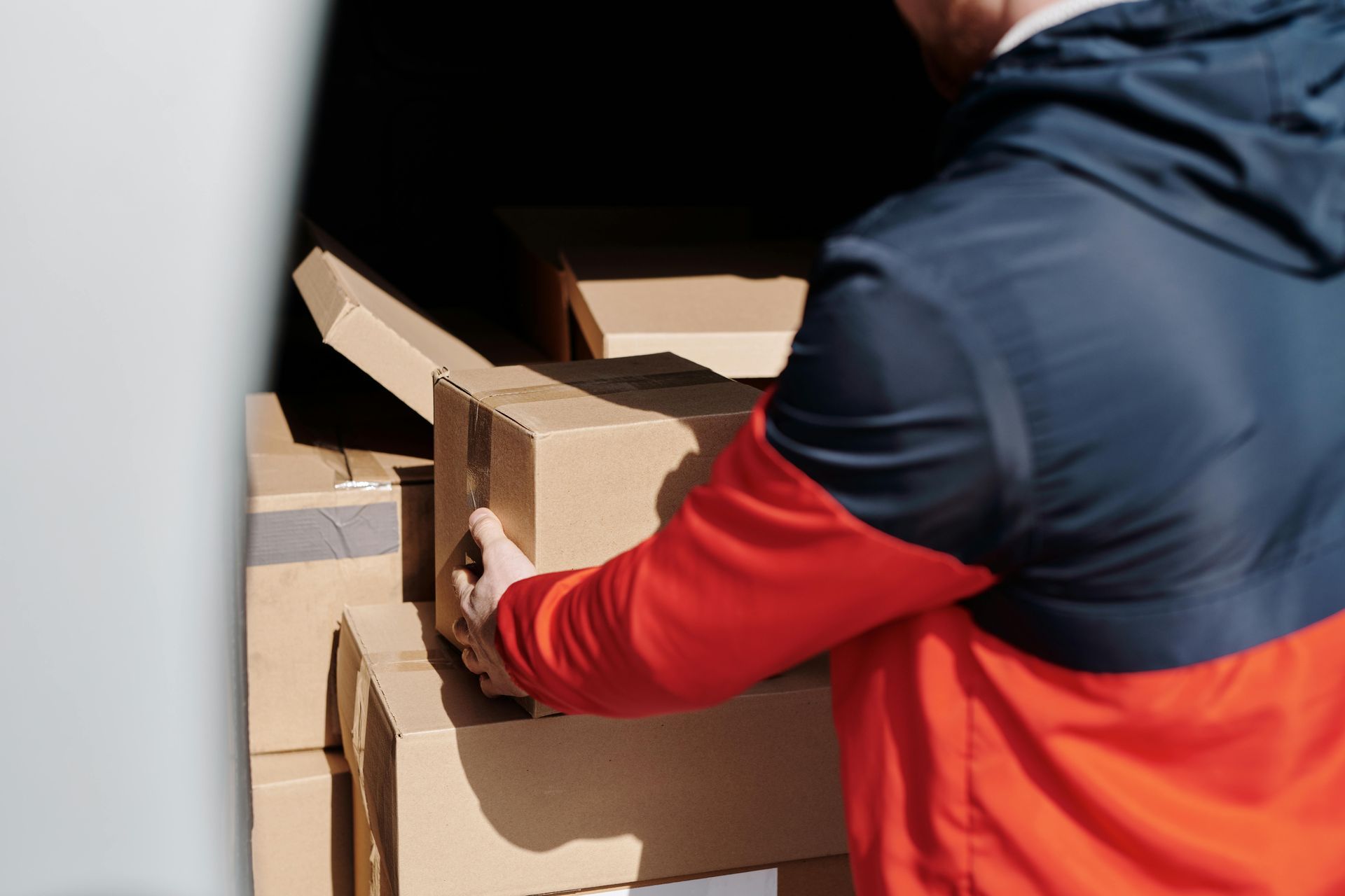 A person wearing a red and navy jacket reaches into a dark vehicle to organize brown cardboard shipping boxes.