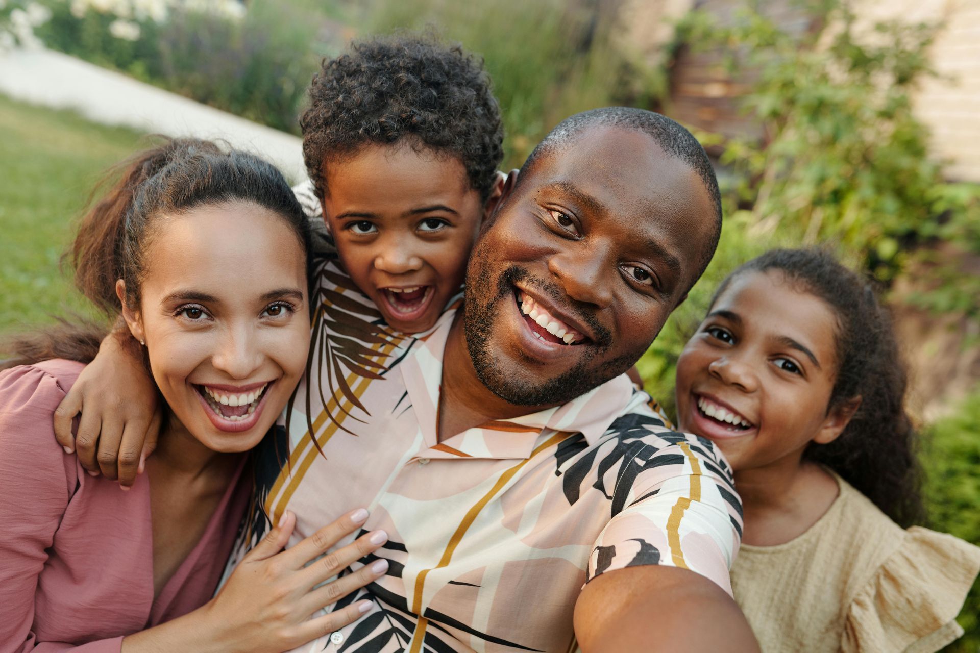 A smiling family of four takes a joyful selfie outdoors in a sunny, green setting.