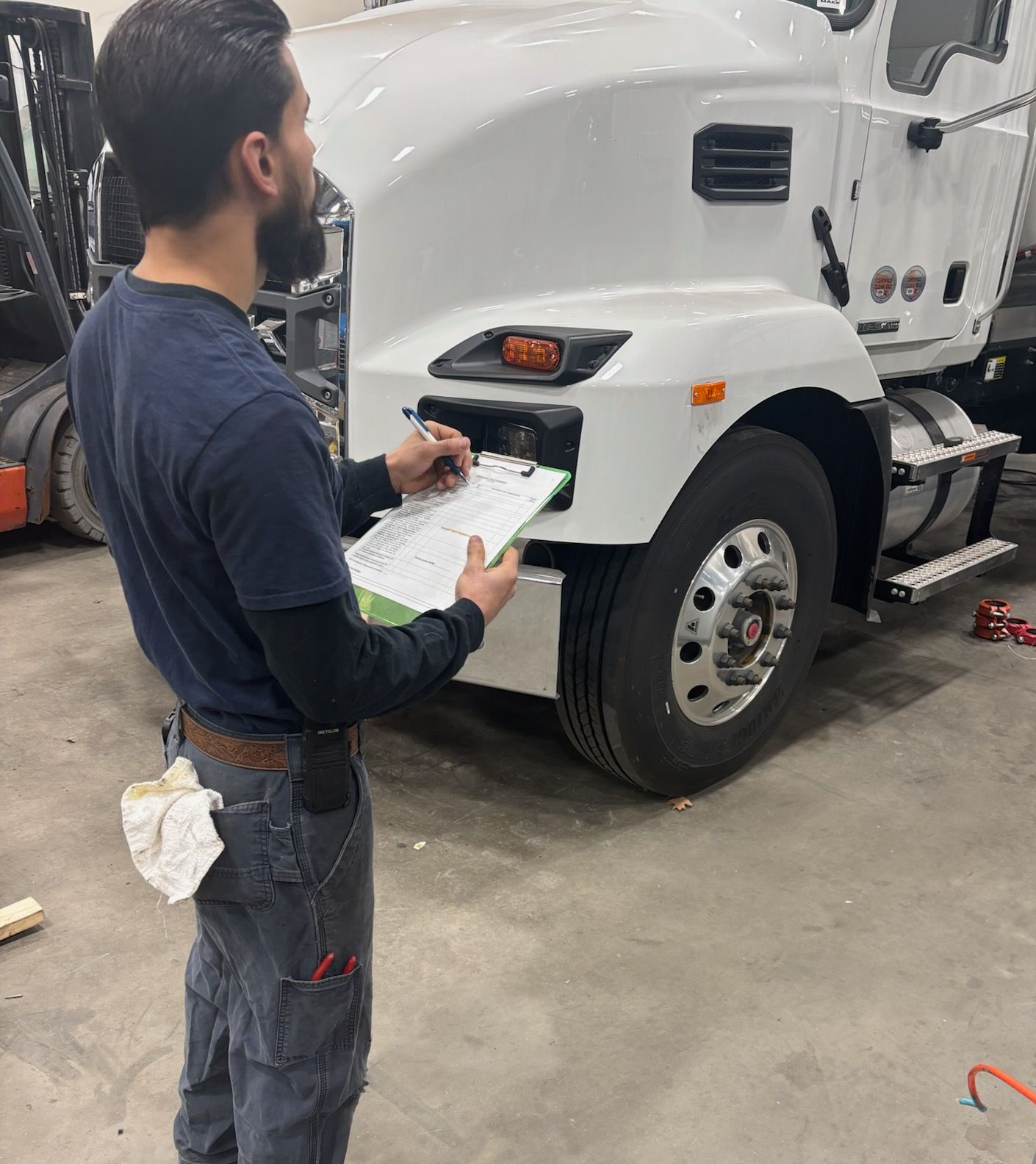 A mechanic in dark work clothes holds a clipboard while inspecting the front of a white semi-truck in a garage.