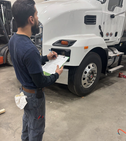 A mechanic in dark work clothes holds a clipboard while inspecting the front of a white semi-truck in a garage.