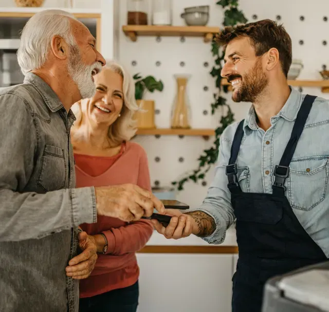 A person in work overalls holds a payment terminal while a couple smiles and prepares to pay in a kitchen.