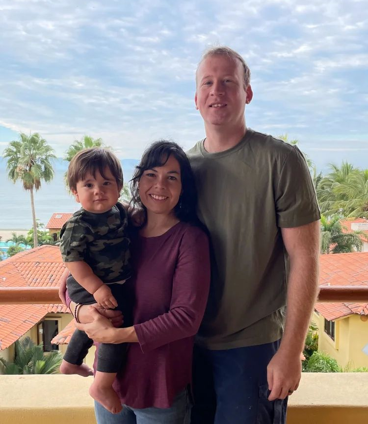 A family of three standing on a balcony overlooking a tropical beach and resort buildings on a sunny day.