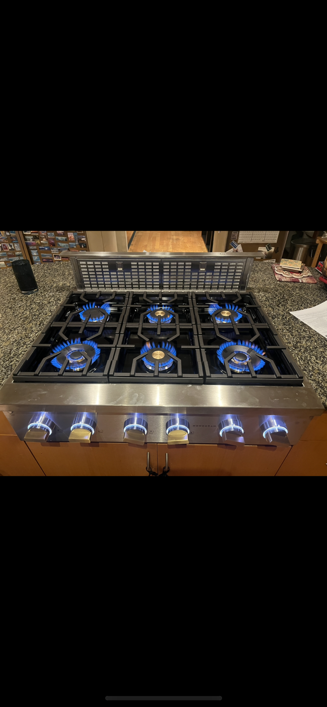 A stainless steel 6-burner gas cooktop with blue flames lit and illuminated control knobs on a granite countertop.