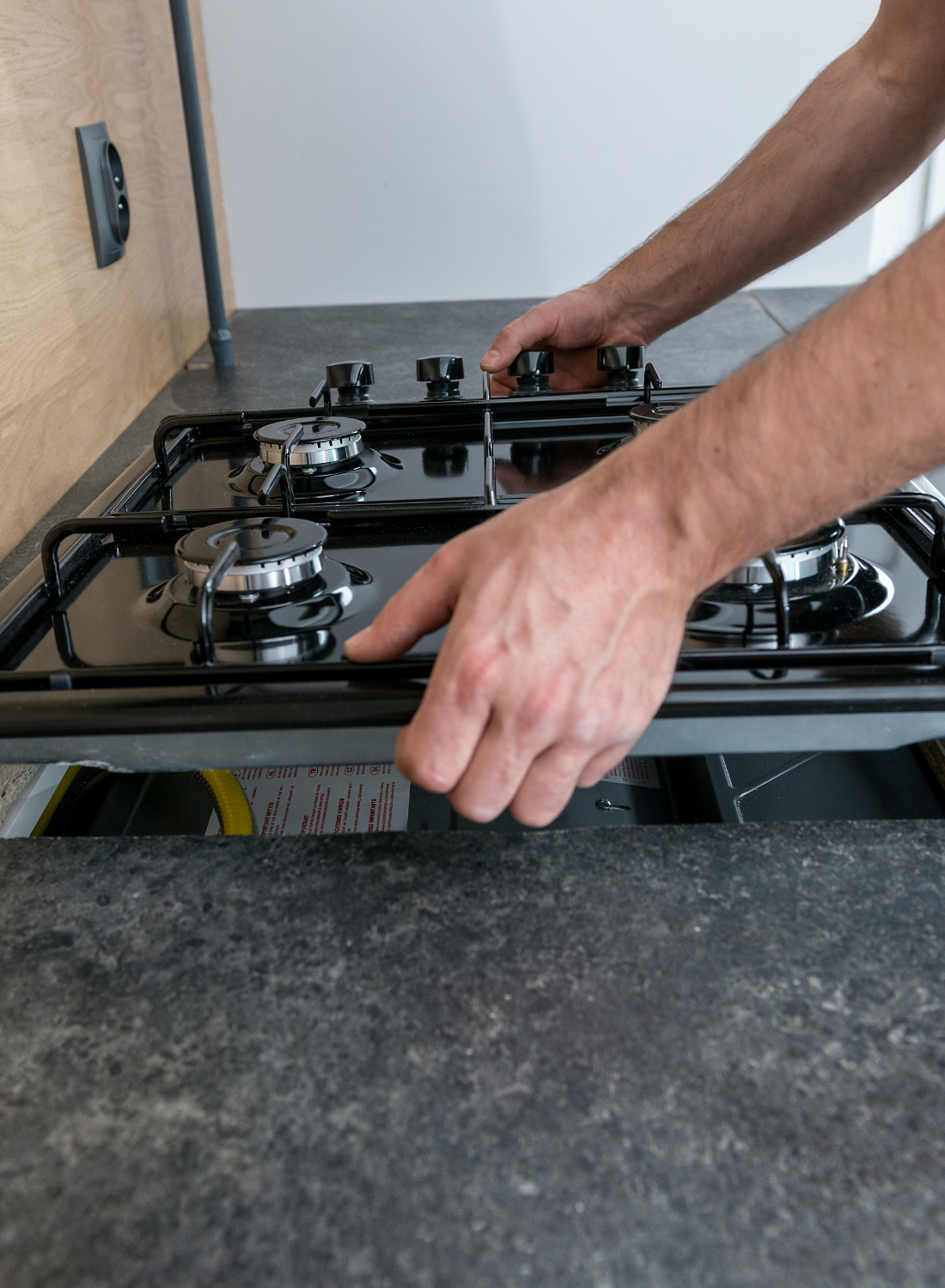 A person lifting a black gas stovetop from a cutout in a dark stone kitchen countertop.