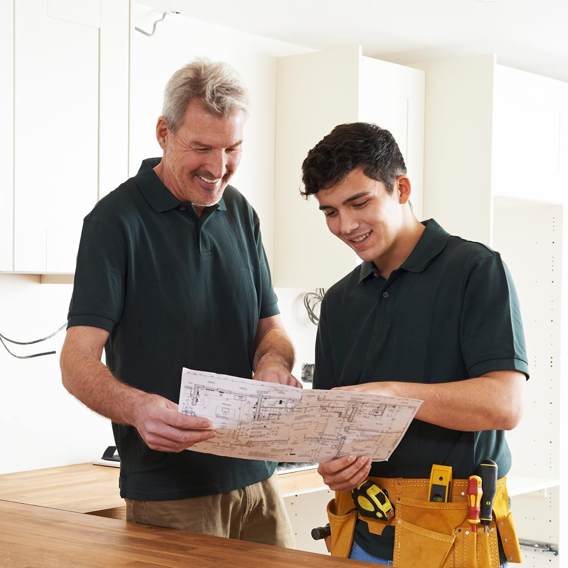 Two workers in green polo shirts look at a blueprint in a kitchen under construction, wearing tool belts.