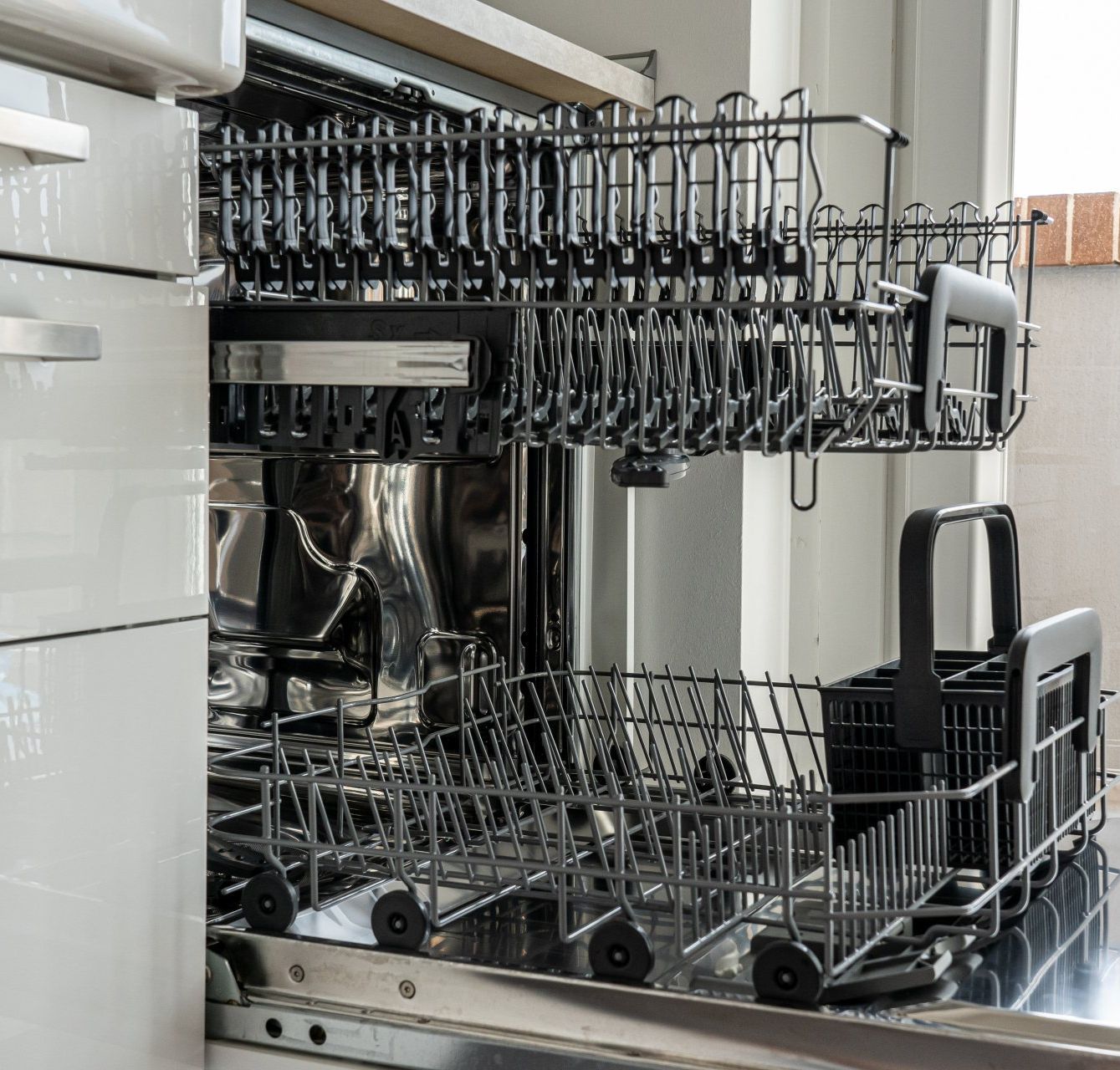 An open, empty dishwasher with two pull-out grey metal racks, featuring a black utensil basket in the lower rack.