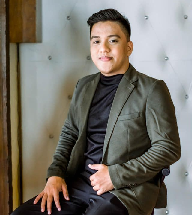 A person in a black mock-neck shirt and olive green blazer sitting against a white tufted backdrop.