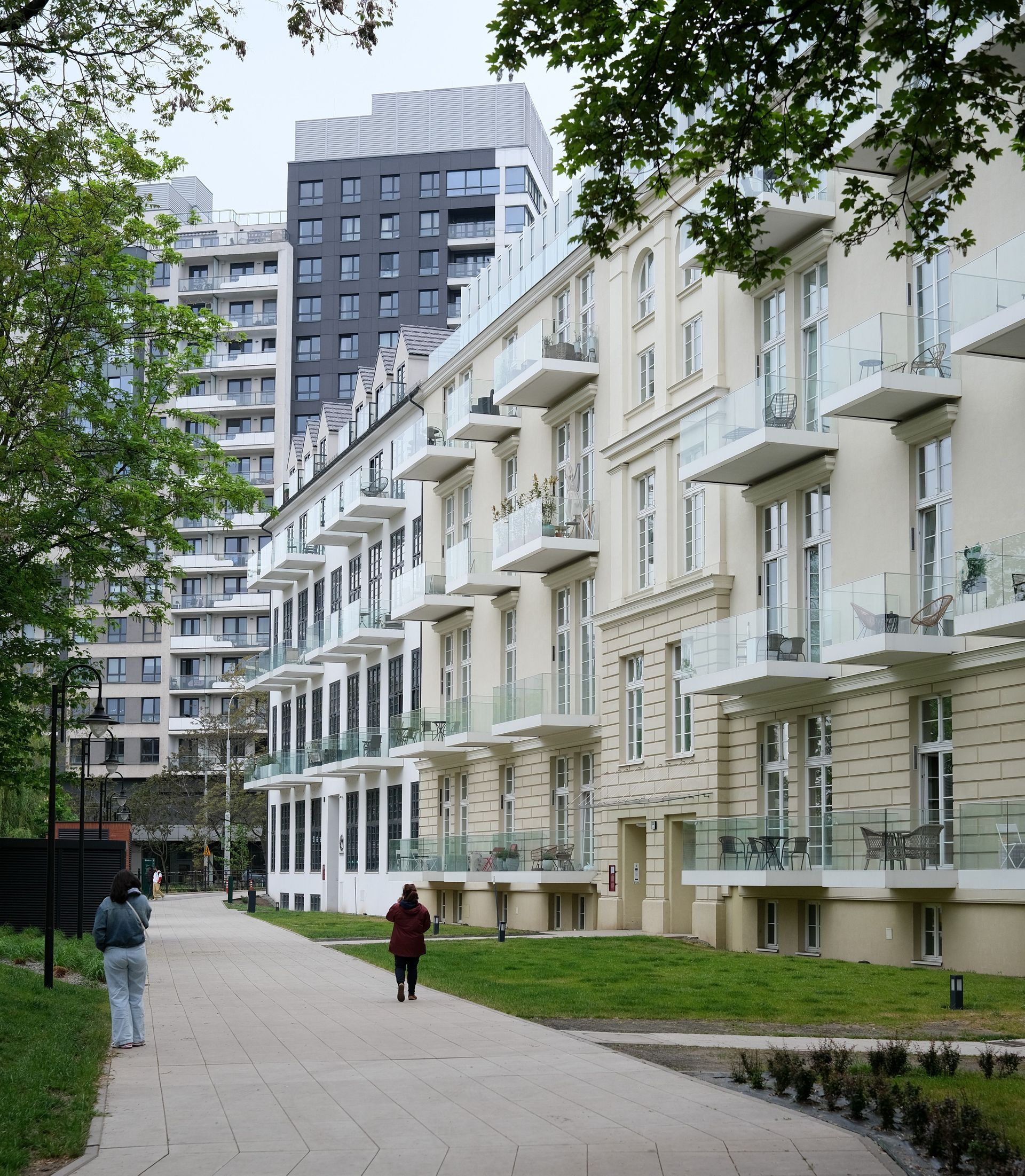 Tree-lined path beside white apartment buildings with balconies and a tall gray tower in the background
