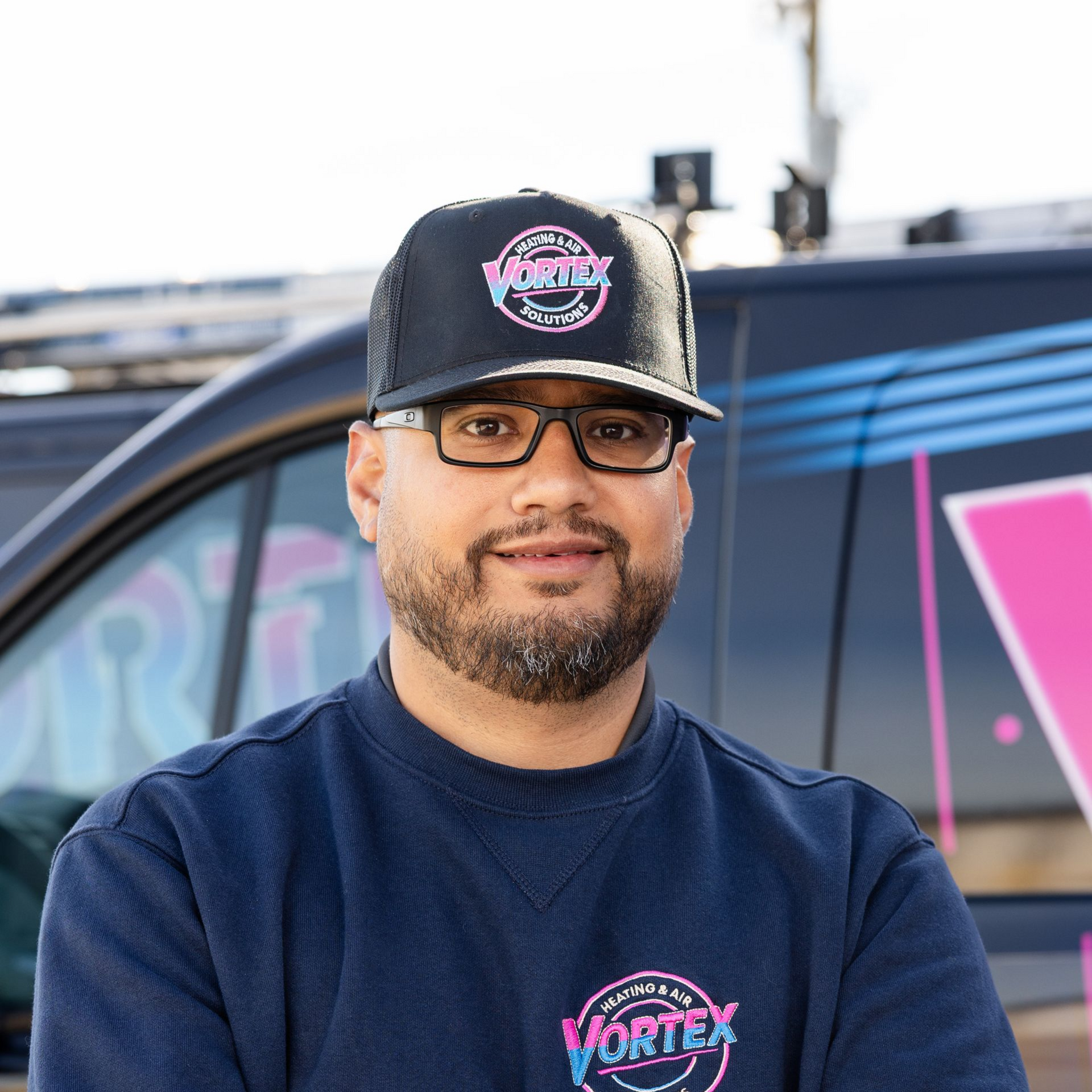Man in black shirt and khaki pants stands in front of a company van with arms crossed.