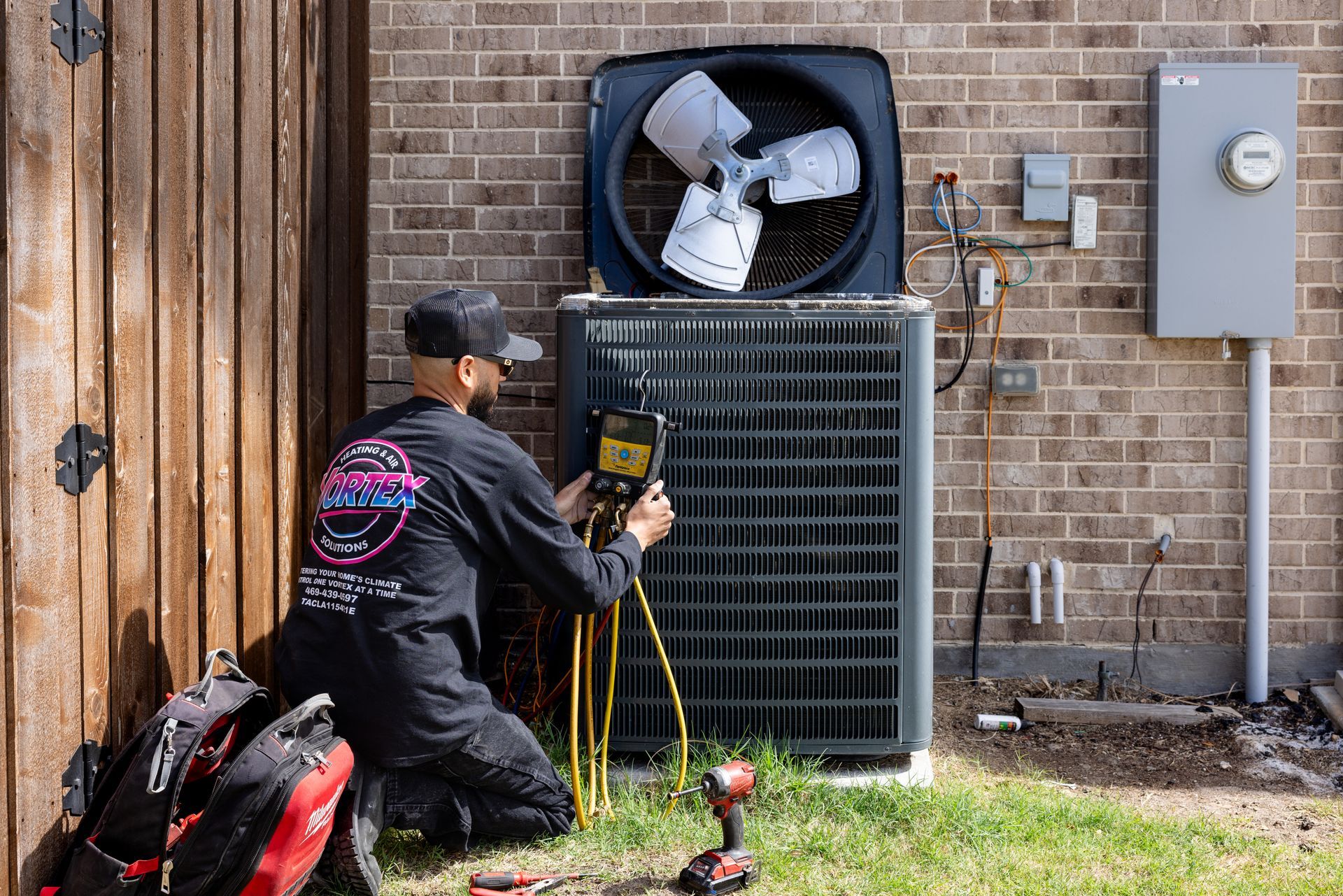 Man in a black shirt working on an AC unit outdoors. He wears a white cap and light blue jeans.