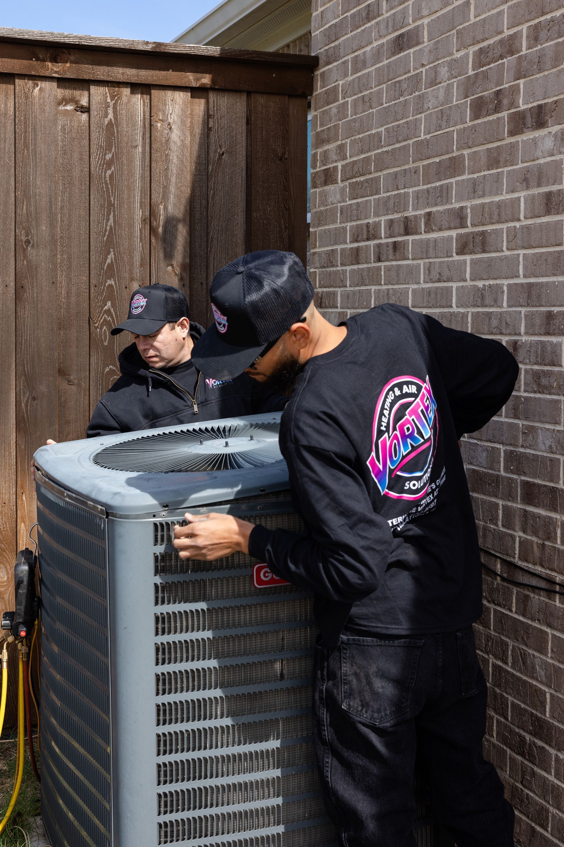 Two men moving a large cardboard box on a hand truck next to a brick house and an air conditioning unit.