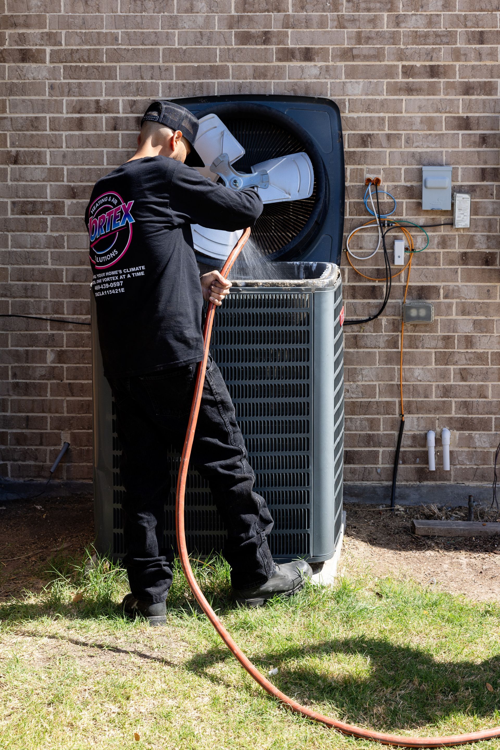 Man cleaning an AC unit with a hose. Gray unit, green hose, wearing a hat, outside near a brick wall.