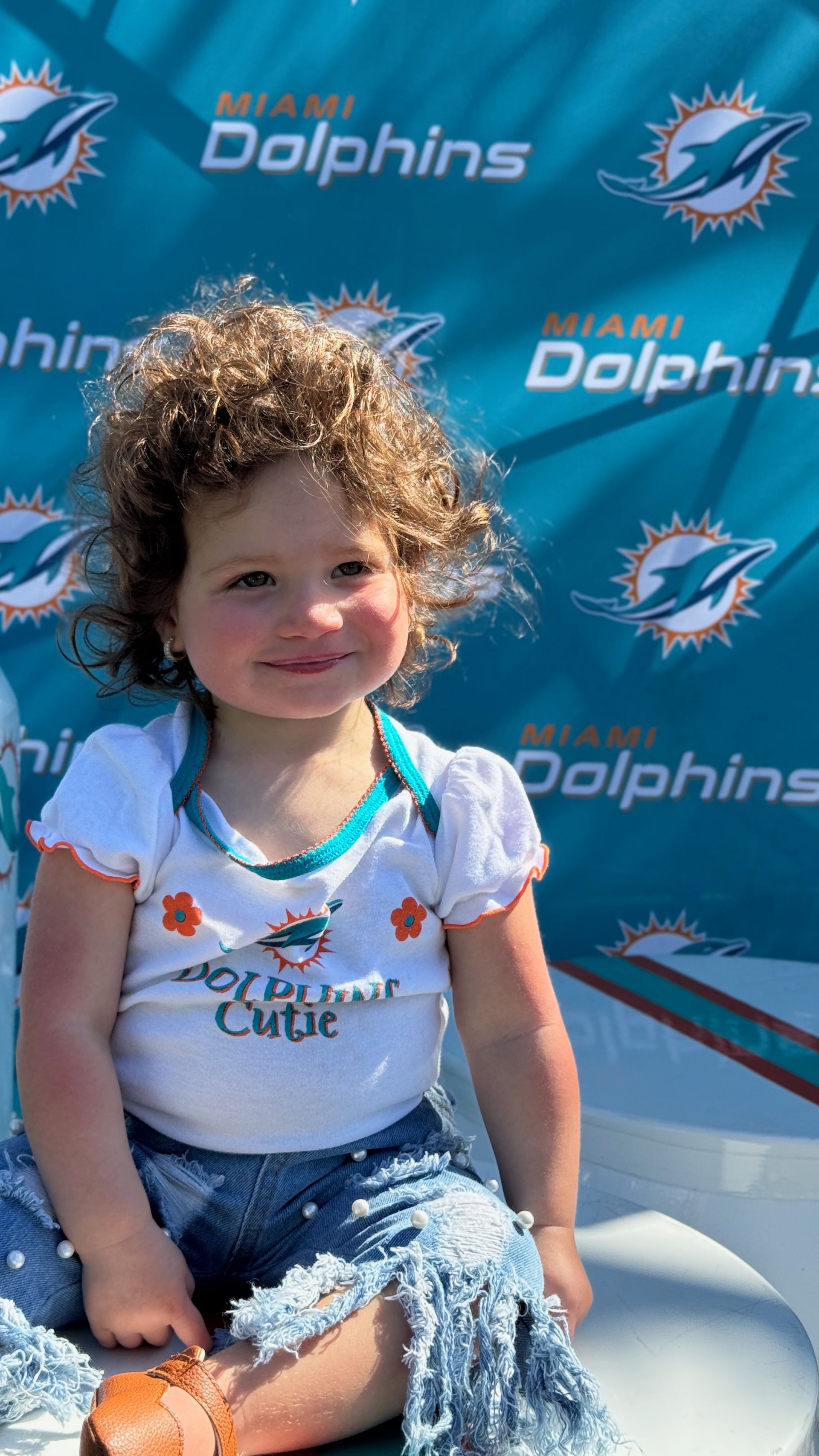A little girl is sitting on a boat in front of a dolphins banner.