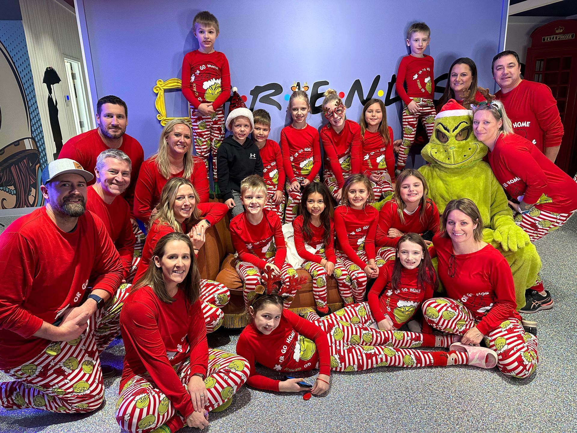 Group of people in red Grinch pajamas and the Grinch pose for a photo in front of a Friends backdrop.