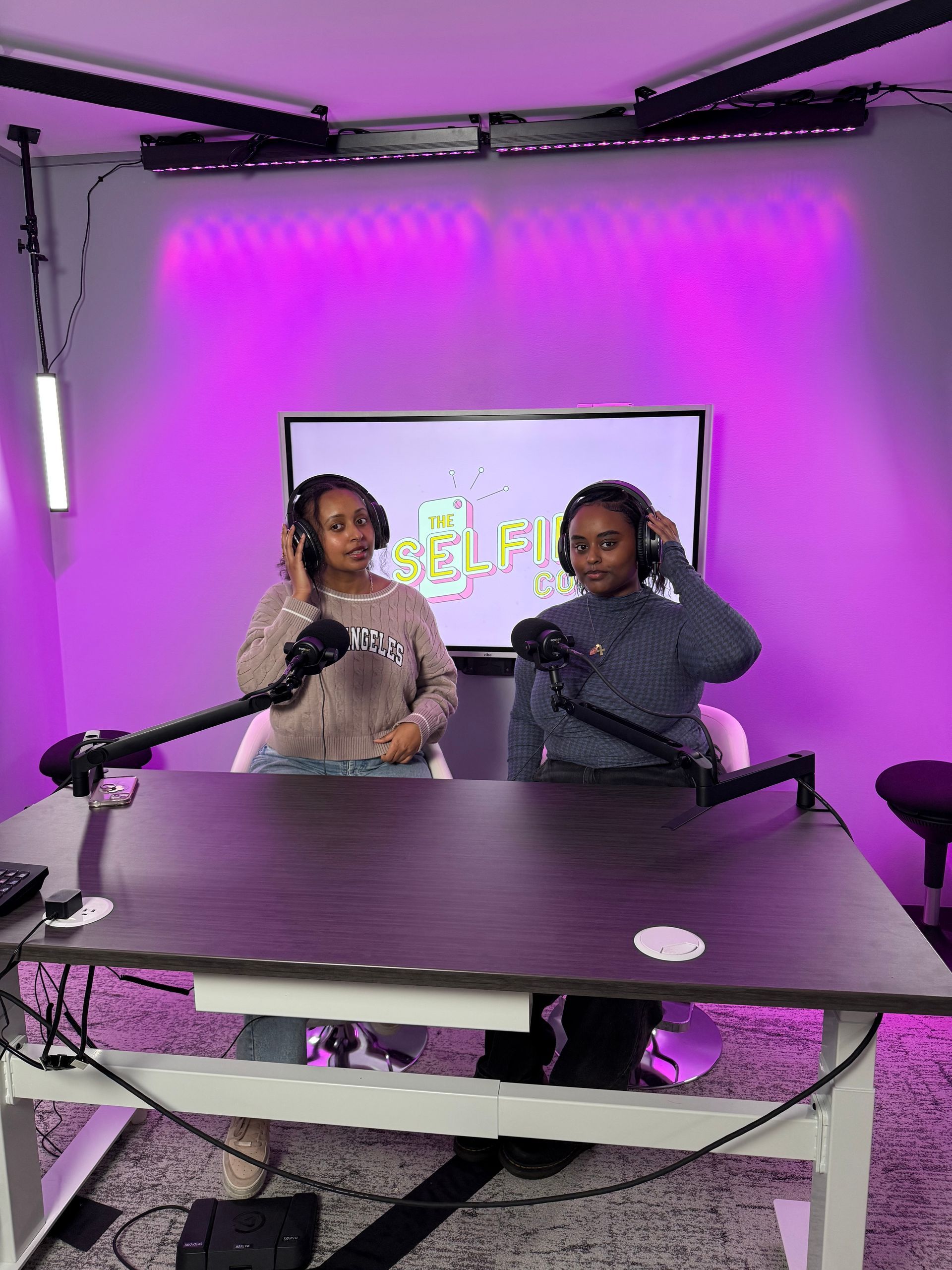 Two people at a podcast desk in a purple-lit studio, with microphones and a screen behind them.