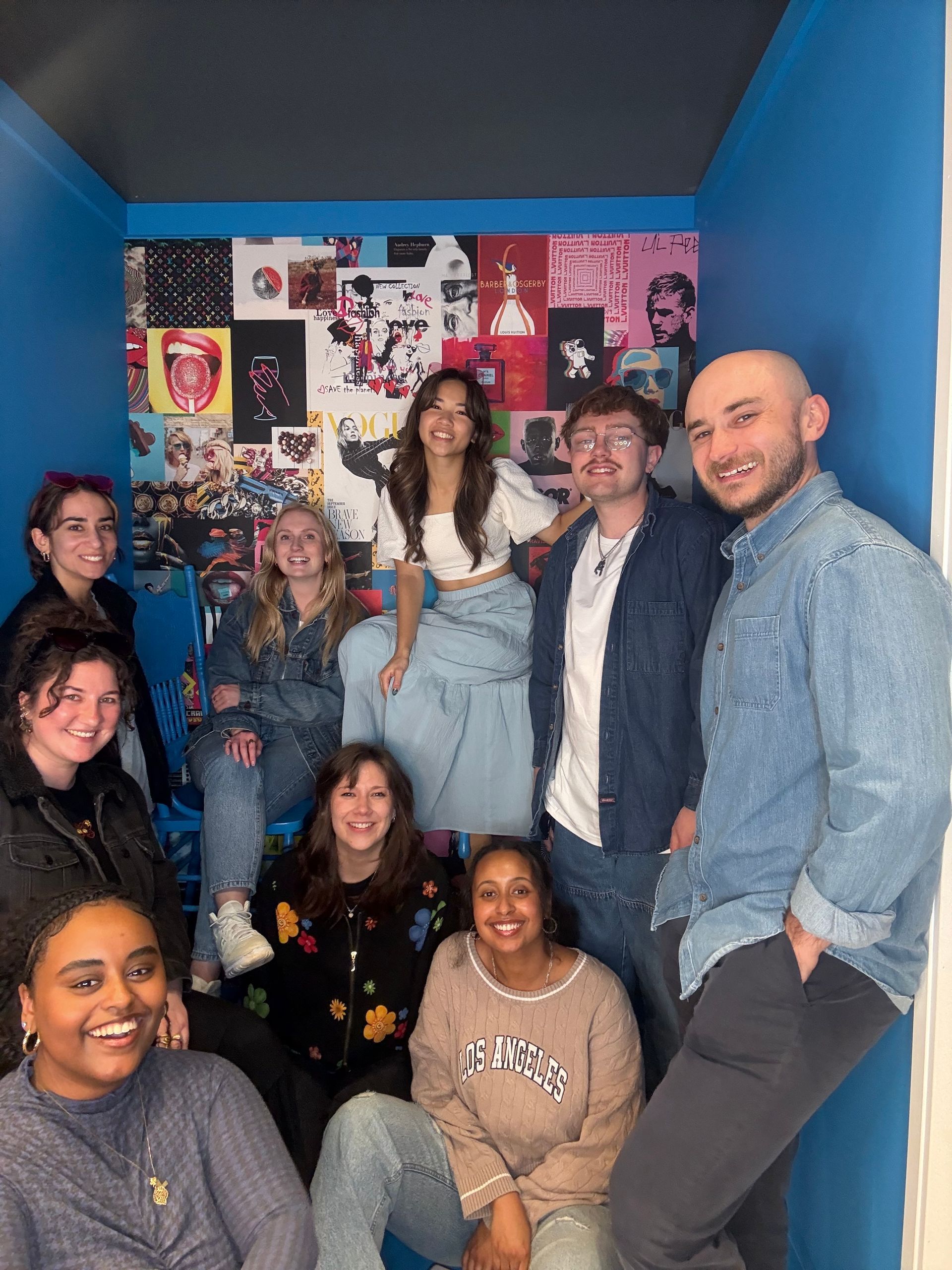 Group of seven smiling people in a blue room with a colorful collage wall.