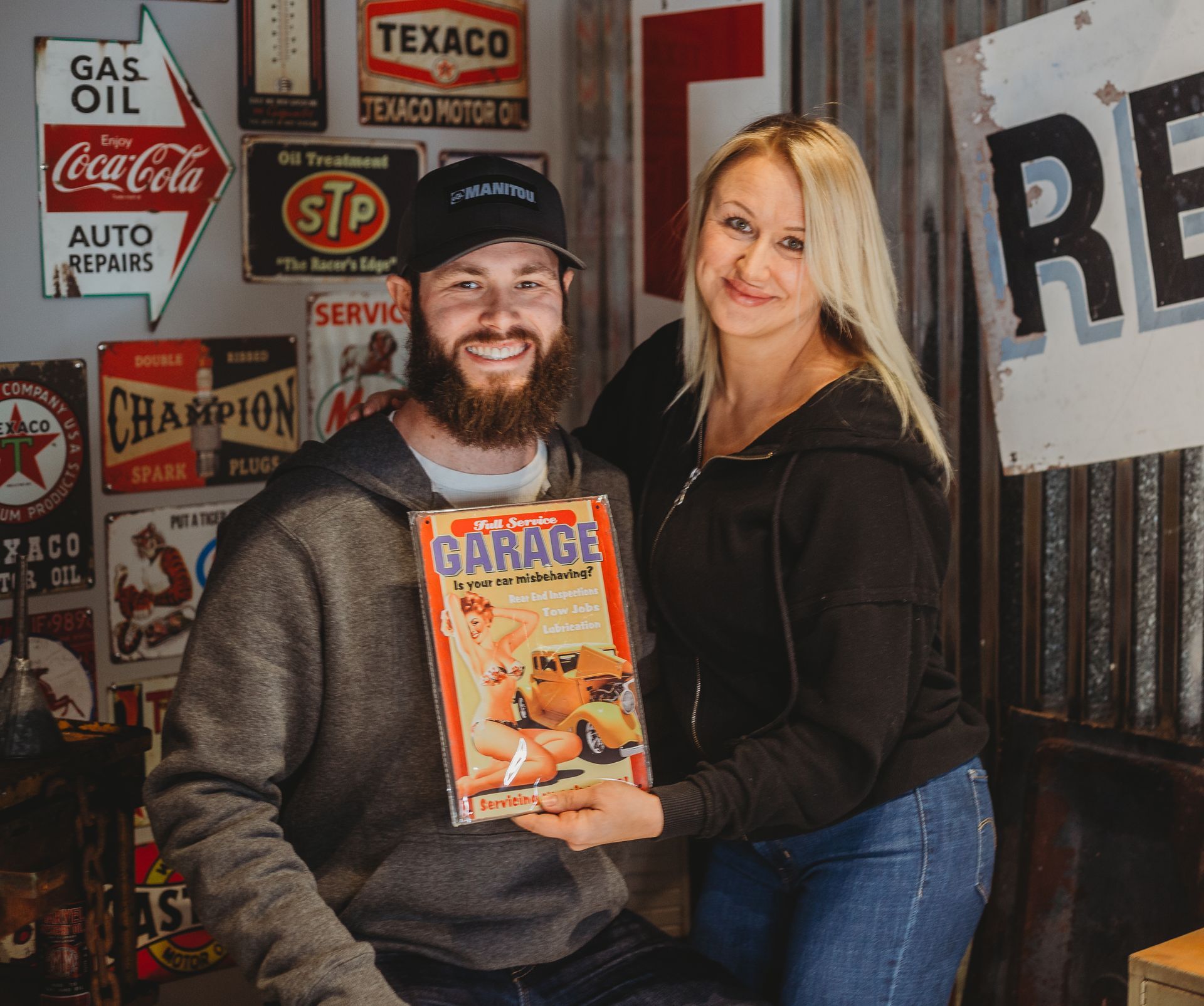 Two people sit in a garage-themed room, smiling and holding a vintage garage sign.
