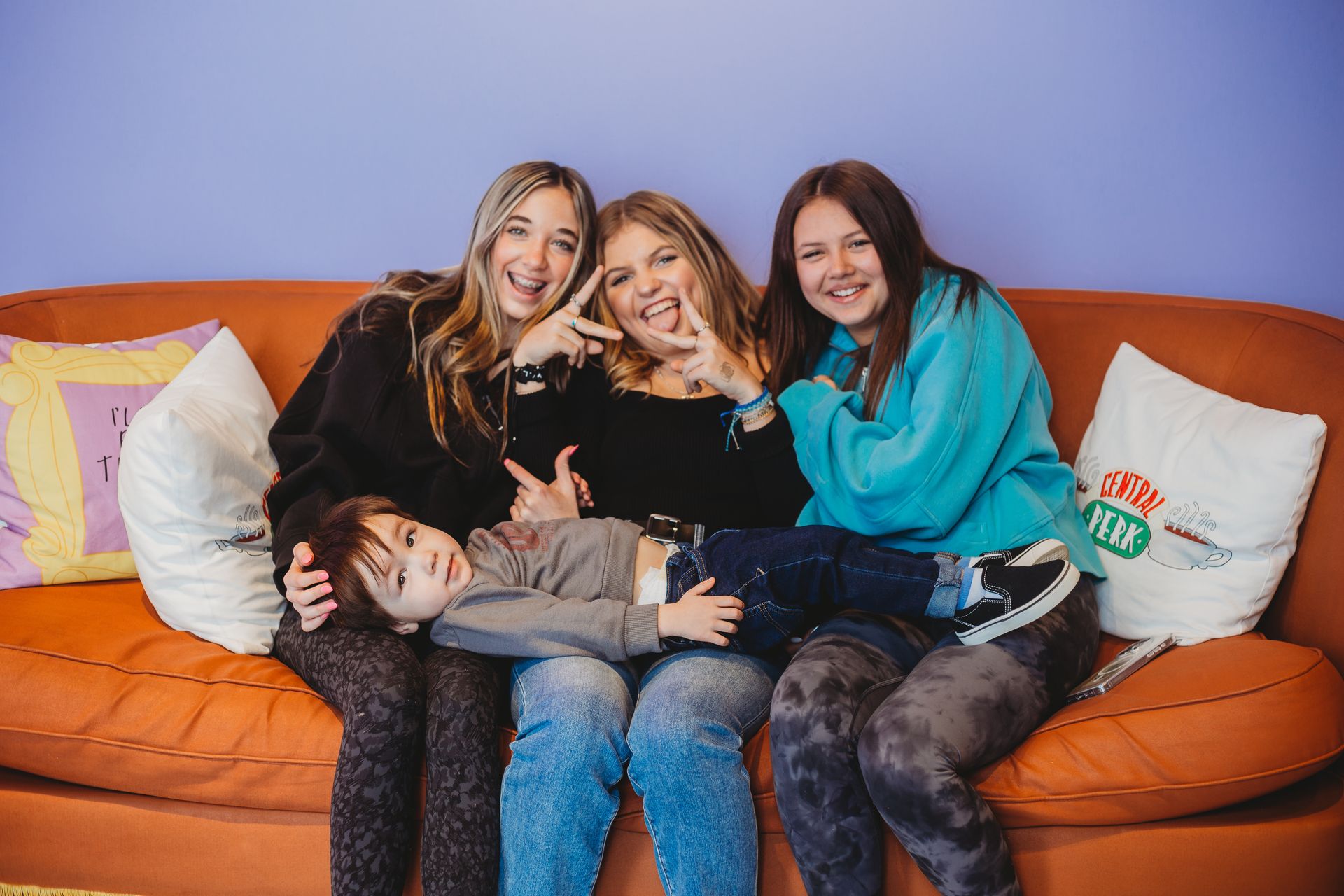 Four friends relaxing on an orange couch, smiling and hugging in a colorful room