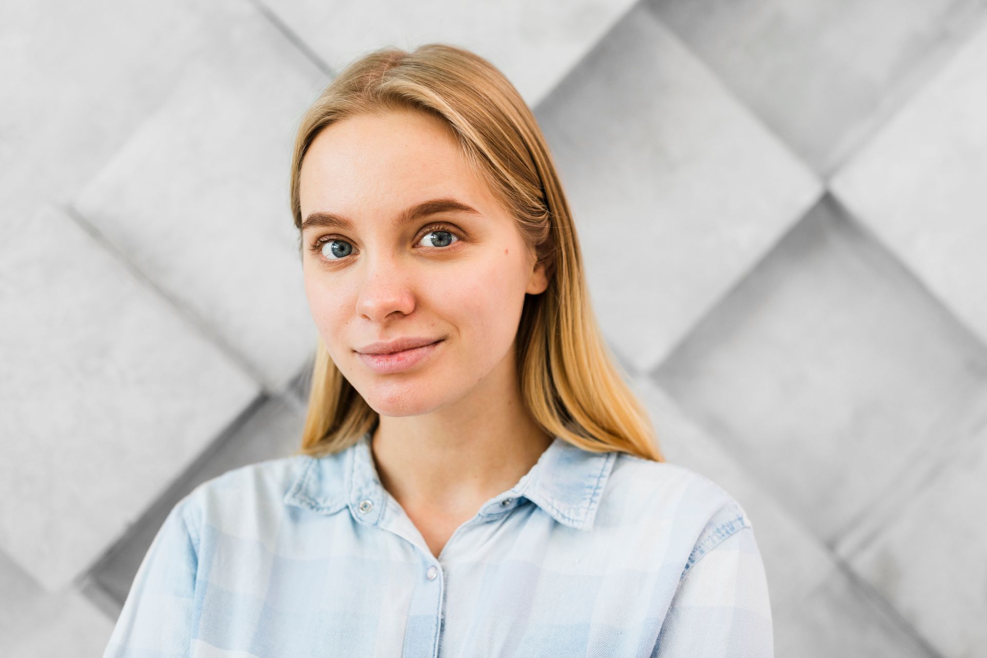 A young woman in a blue shirt is standing in front of a gray wall.