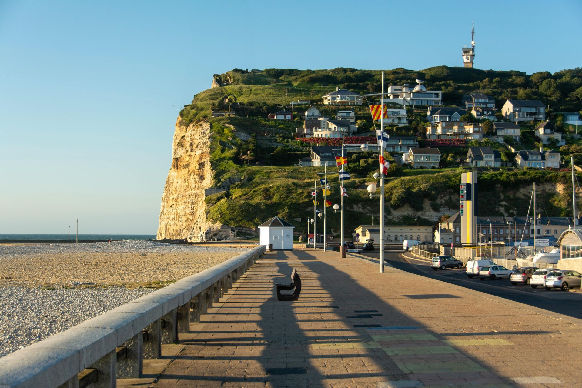 Promenade côtière longeant une plage de galets et des maisons perchées sur les falaises au coucher du soleil