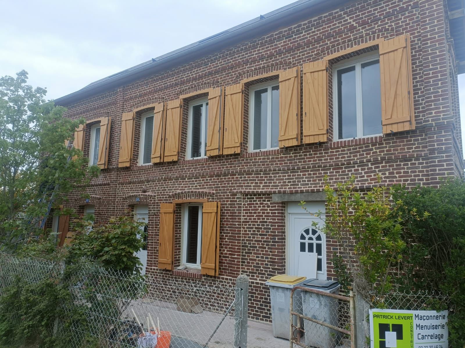 Maison en briques de deux étages avec volets beiges, porte blanche et cour clôturée envahie par la végétation.