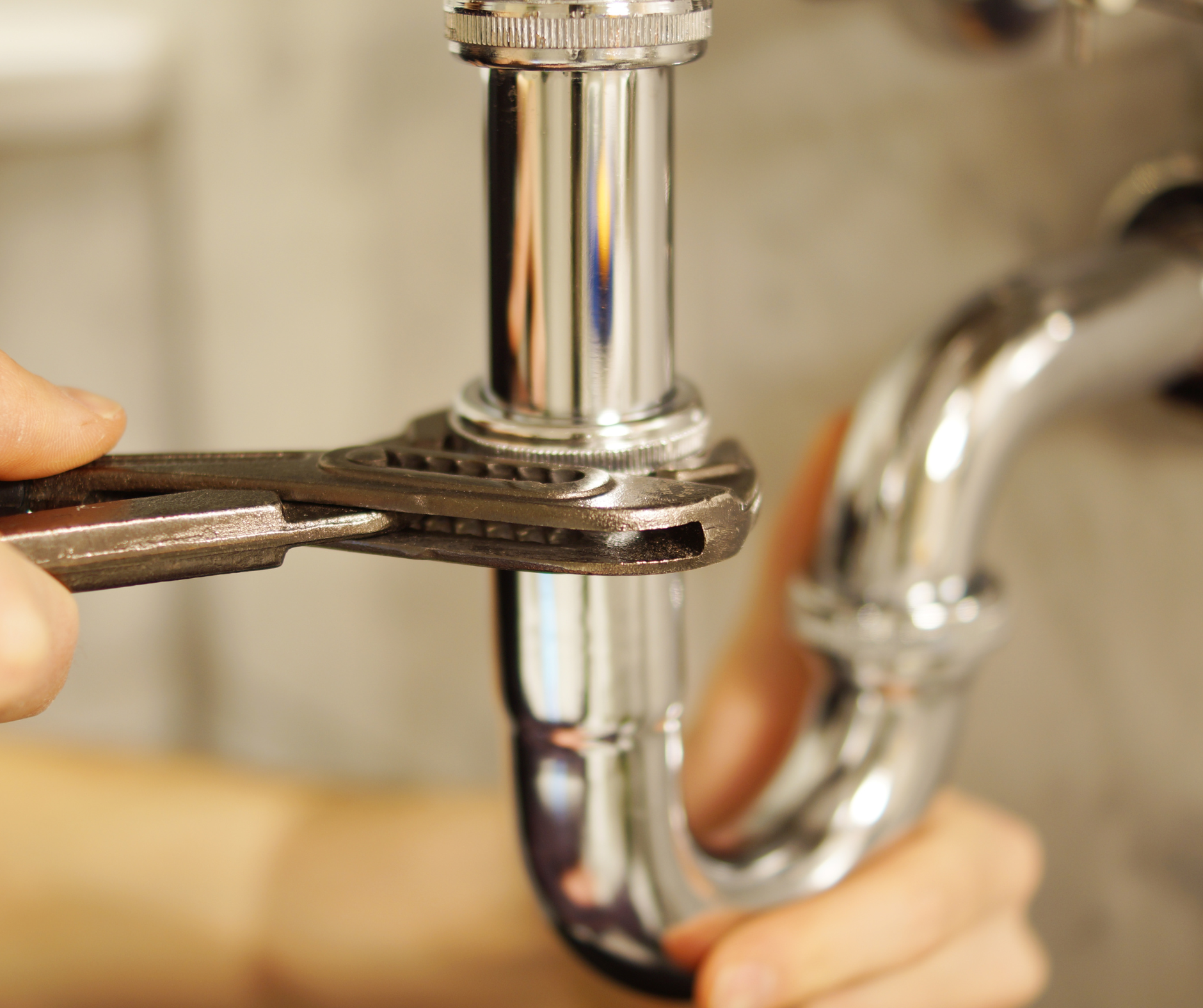 A person using a wrench to tighten a chrome pipe under a sink.