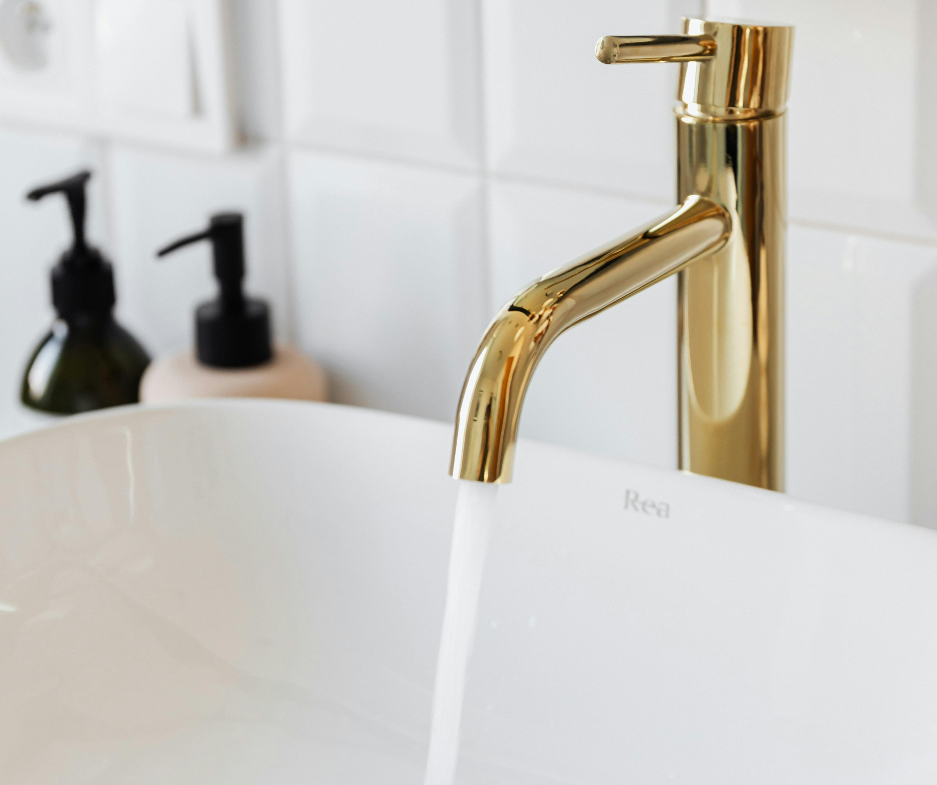 Gold faucet pouring water into a white sink, soap dispensers in the background.