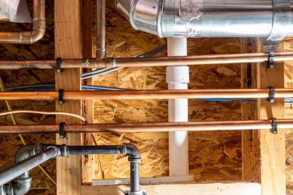 Plumbing pipes: copper, black, and white PVC against a wood-paneled wall, held by black clips.