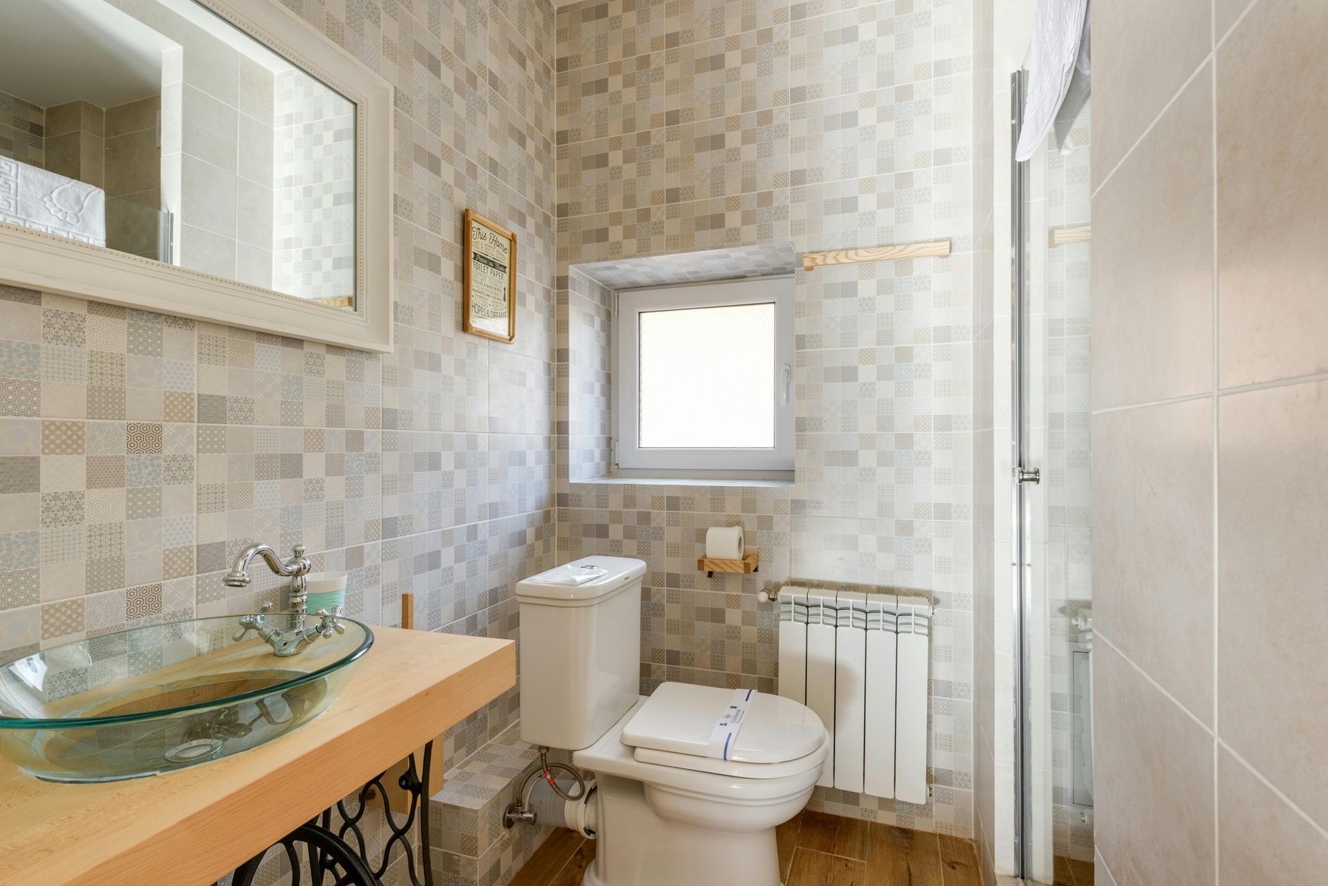 Small bathroom with mosaic tile walls, glass sink, toilet, small window, and shower door.
