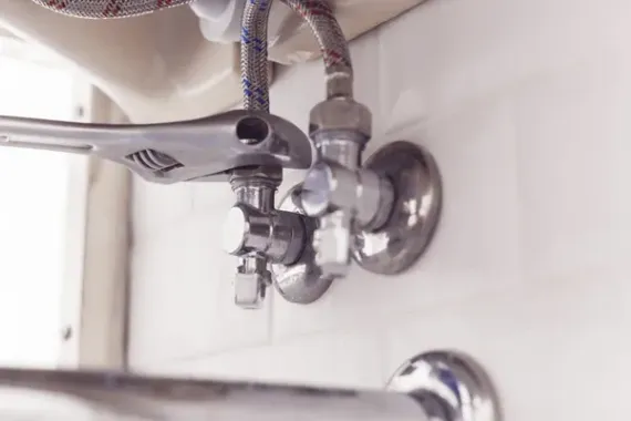 A wrench tightening a faucet valve in a white-tiled bathroom, connected to flexible water supply lines.