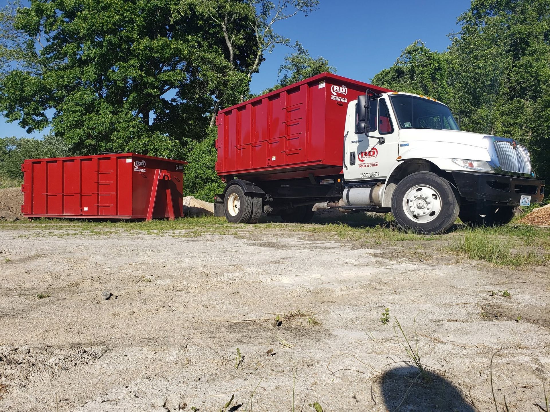 Red dump truck beside a large red dumpster on a gravel lot, with trees in the background
