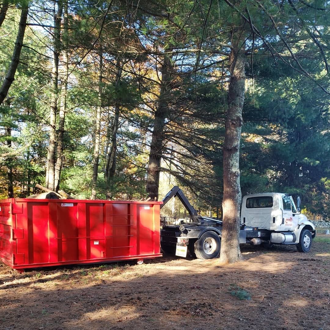 Red dumpster beside a small white utility truck in a wooded area with tall pine trees.