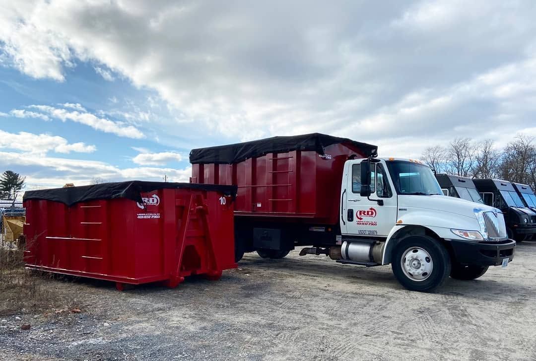 Red dump truck with a red dumpster trailer parked on a gravel lot under a cloudy sky