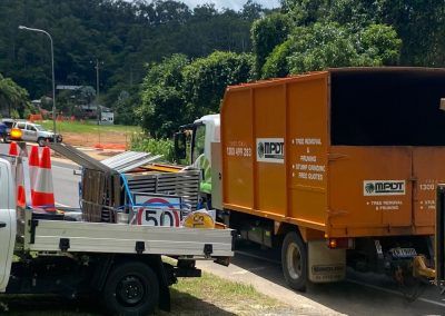 An orange dumpster truck and white pickup truck at a roadside work site. 50km/h speed limit sign visible.