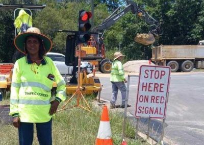 Traffic control workers at road construction site. One standing by a 'stop here' sign. Others near machinery and traffic lights.